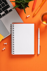 Blank notebook with laptop keyboard, pens, and cup of tea on orange desk. Workspace for study,...