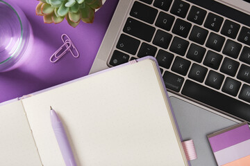 Notebook with pen near laptop keyboard, plant, and glass of water on purple desk. Place for your...