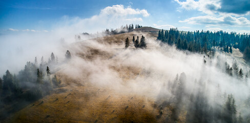Stunning landscape with dense forests and rolling hills partially shrouded in soft, ethereal fog. Vibrant autumn colors of trees contrast beautifully with clear blue sky and distant mountain peaks.