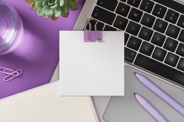 Blank note with binder clip on laptop keyboard with pens, notebook, and plant on purple desk....