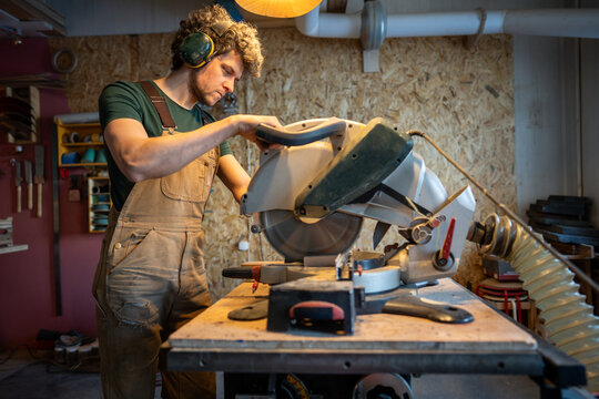 Skilled focused carpenter man in earmuffs use miter saw cut wood beams in joinery. Handyman precision cutting, woodworking craftsmanship, blade control, furniture making business. 