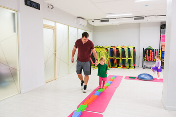 A man holding a girl's hand as they walk on balance stepping stones on a pink mat in a gym during an exercise session.