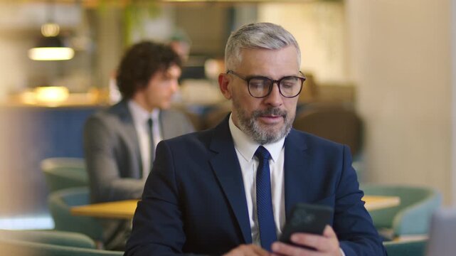 Middle aged Caucasian businessman in glasses and formal suit checking his smartphone and smiling while sitting in stylish cafe