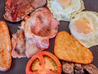 English breakfast with soft eggs, potato cake, mushrooms, back bacon, link sausage, grilled tomato, and a small beef steak on a black plate