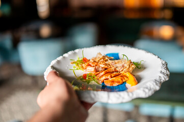 Close-up of gourmet shrimp dish on ornate white plate, served with yellow puree and microgreens in upscale restaurant setting, held by hand. Fine dining presentation