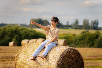 A smiling boy sits barefoot on a hay bale in a golden field, pointing joyfully into the distance on a sunny day, surrounded by countryside scenery.