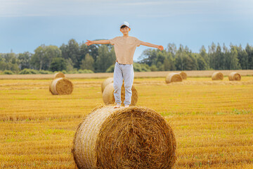 Smiling boy in a cap stands barefoot on a hay bale with arms outstretched, enjoying the golden...