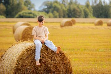 Smiling boy sitting barefoot on a round hay bale in a golden field with more bales scattered in the...