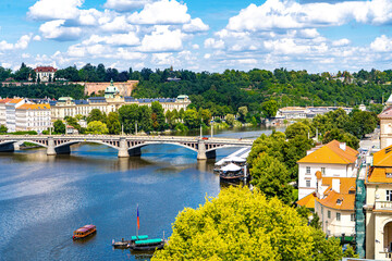 Obraz premium he Vltava River, surrounded by lush summer greenery, with a bridge connecting the historic districts of the Old Town under a blue sky with clouds.Prague,Czech Republic