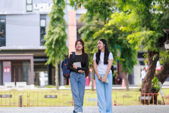 Two asian female students carrying backpacks and holding tablet and book are walking and talking together in university campus