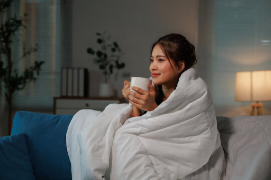 Asian woman sitting on comfortable sofa covered with warm blanket holding a cup of hot beverage looking away and smiling in cozy living room at night