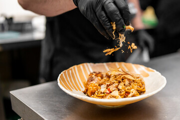 Chef garnishing noodle dish with almond flakes in professional kitchen. Close-up of gloved hand, striped bowl, and stainless steel counter. Culinary plating and food styling.