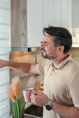 Man drinking coffee and opening blinds in a modern kitchen