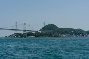 Coastal bridge spans over the blue ocean, connecting a green, hilly island with transmission towers visible.