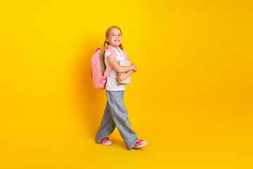 Charming young schoolgirl with a pink bag on a bright yellow background holding a book walking confidently and smiling.