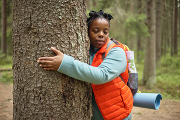 Black woman hugging tree in forest, wearing backpack and carrying rolled yoga mat, eyes closed,...