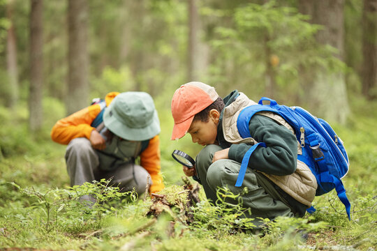 Two children exploring forest floor, one boy with blue backpack using magnifying glass to examine plants while another child in background observing nature closely