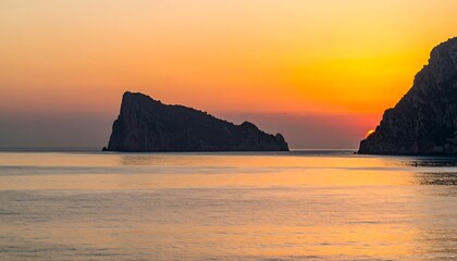 Fototapeta premium Serene sunset over calm ocean, two dark silhouettes of rocky islands stand in the foreground, reflecting the warm light