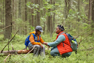 Black woman supporting Black girl while sitting together in forest, both wearing backpacks, woman...
