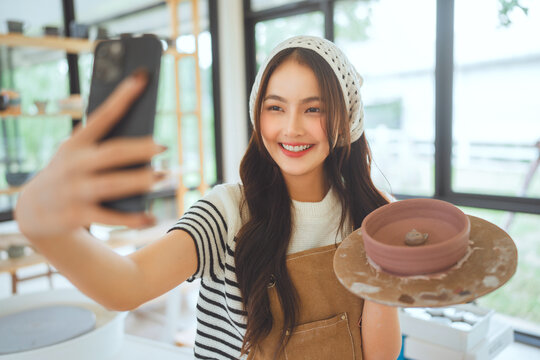 Happy asian female artist taking selfie with handmade pottery design in creative studio – smiling sculptor sharing artwork on social media for online selling, digital portfolio and creative expression - Powered by Adobe