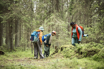 Obraz premium Mother and kids hiking in forest, standing on trail and observing ground closely, carrying backpacks and outdoor gear, surrounded by dense green trees