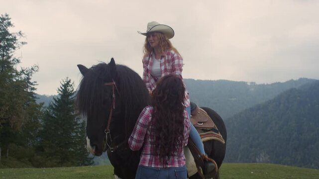 Young woman at a western riding school in a stunning mountain setting, mounting the horse while holding onto the saddle, assisted by a cowgirl instructor, handheld shot.