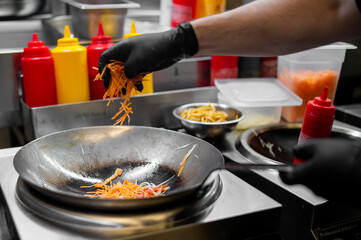 Chef cooking stir-fry in commercial kitchen, adding shredded vegetables to wok. Professional food preparation with sauces, bowls, and fresh ingredients in background