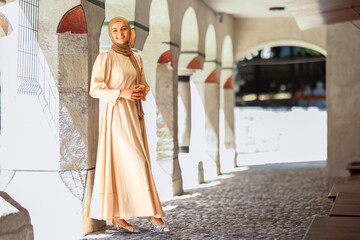 Fototapeta premium Muslim woman in modest beige dress and hijab smiling under traditional stone arches, cultural heritage and elegant fashion in historic architecture.