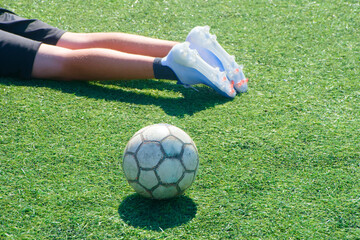 Worn soccer ball on green artificial turf near the legs of a young player in cleats, game paused with a hint of injury during a youth match in sun. Photo