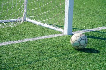 Old soccer ball by the goalpost on green artificial turf, right on the goal line — a tense near‑miss at the corner of the net in bright sun. Photo