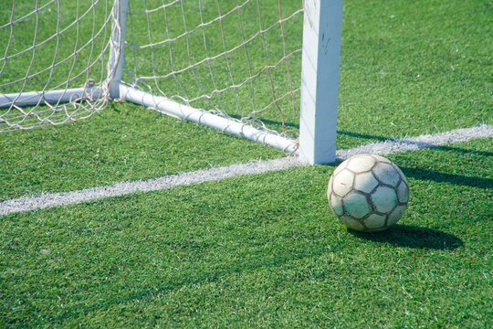 Old soccer ball by the goalpost on green artificial turf, right on the goal line &mdash; a tense near‑miss at the corner of the net in bright sun. Photo
