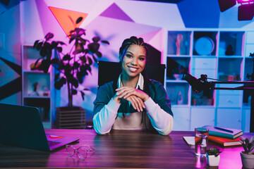Charming female podcaster sitting at a desk with a microphone and laptop in neon lighting for a...