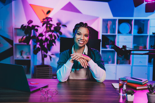 Charming female podcaster sitting at a desk with a microphone and laptop in neon lighting for a media interview