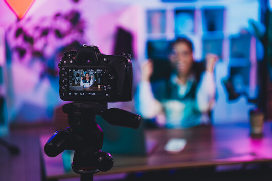 Focused camera capturing young female blogger in colorful studio setting surrounded by equipment for professional content creation