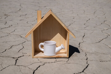 A miniature watering can in a symbolic wooden house on clay cracked by the heat in the desert