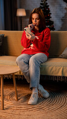 A young woman sits on a sofa and browses on a smartphone. The female is wearing a festive Christmas...