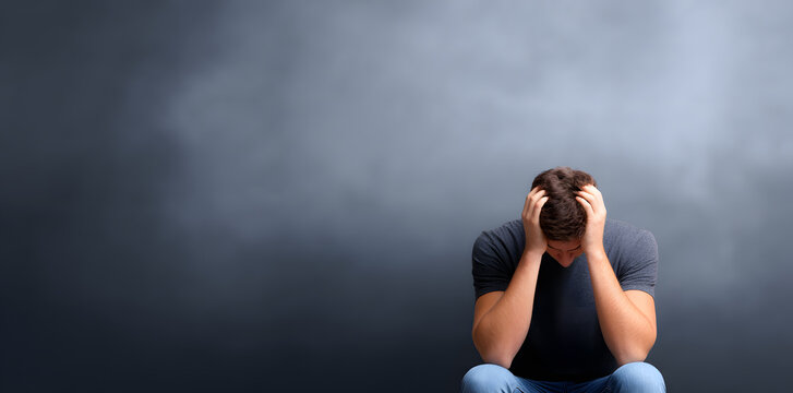 Man sitting against dark wall with head in hands. Concept of hopelessness, burnout, and emotional shutdown in dim environment.