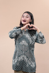 Asian woman in traditional batik shirt smiling brightly while making a heart hand gesture, expressing happiness, love, and positive emotion against plain background.