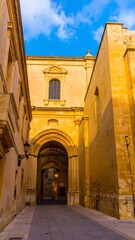 Fototapeta premium A sun-drenched alleyway leads past a historic yellow stone building, showcasing intricate arches and architectural details under a vibrant blue sky.