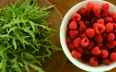 Kitchen board with bunch of fresh arugula and bowl with ripe raspberries on wooden table flat lay stock photo
