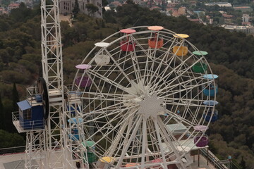 Colorful Ferris wheel with bright cabins in a park, set against a backdrop of trees and city...