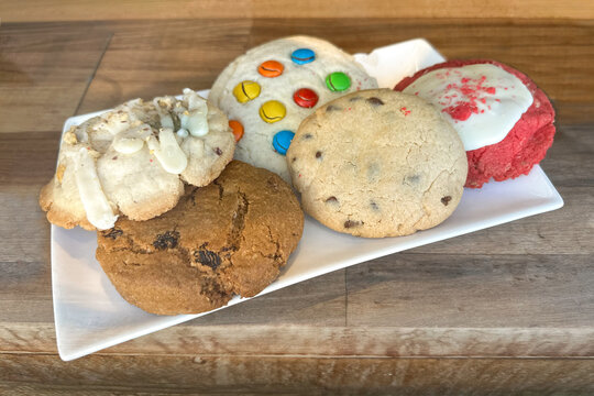 Close-up of assorted cookies on a white plate placed on a rustic wooden table. The variety includes chocolate chip, oatmeal raisin, red velvet with icing, nut cookie with drizzle, and a colorful candy