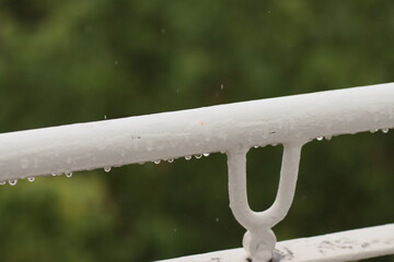 Close-up of a white metal railing with raindrops clinging to its surface, set against a blurred...