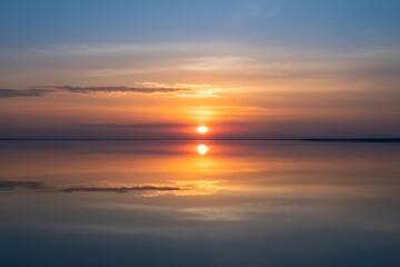 Fototapeta premium Sun Setting Over Calm Water Reflecting Orange and Blue Sky with Clouds sunset setting sun