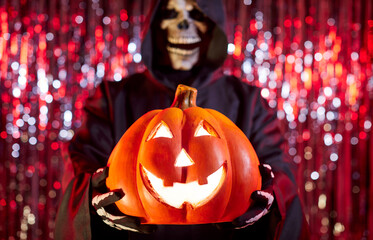 Grim reaper person dressed in mask dark hooded black cloak holds glowing carved pumpkin against a...
