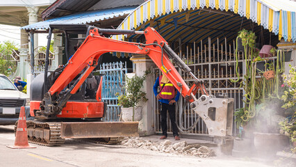 Excavator with crushing breaker is breaking concrete pavement floor in front of residential area to carry out underground water pipe replacement work.