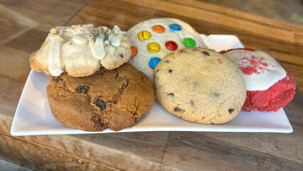 Close-up of assorted cookies on a white plate placed on a rustic wooden table. The variety includes chocolate chip, oatmeal raisin, red velvet with icing, nut cookie with drizzle, and a colorful candy