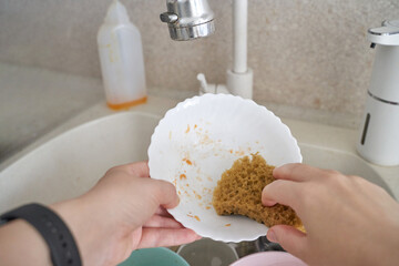 Close up of hands washing dishes in kitchen sink with sponge and soap under running water.