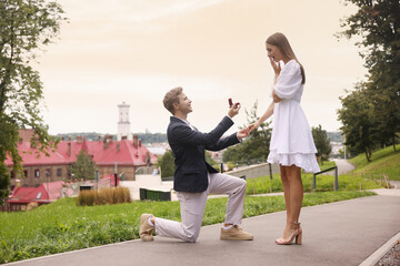 Smiling man with engagement ring making marriage proposal to his surprised girlfriend outdoors....