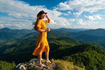 Woman in yellow dress with hat standing on top of mountain.